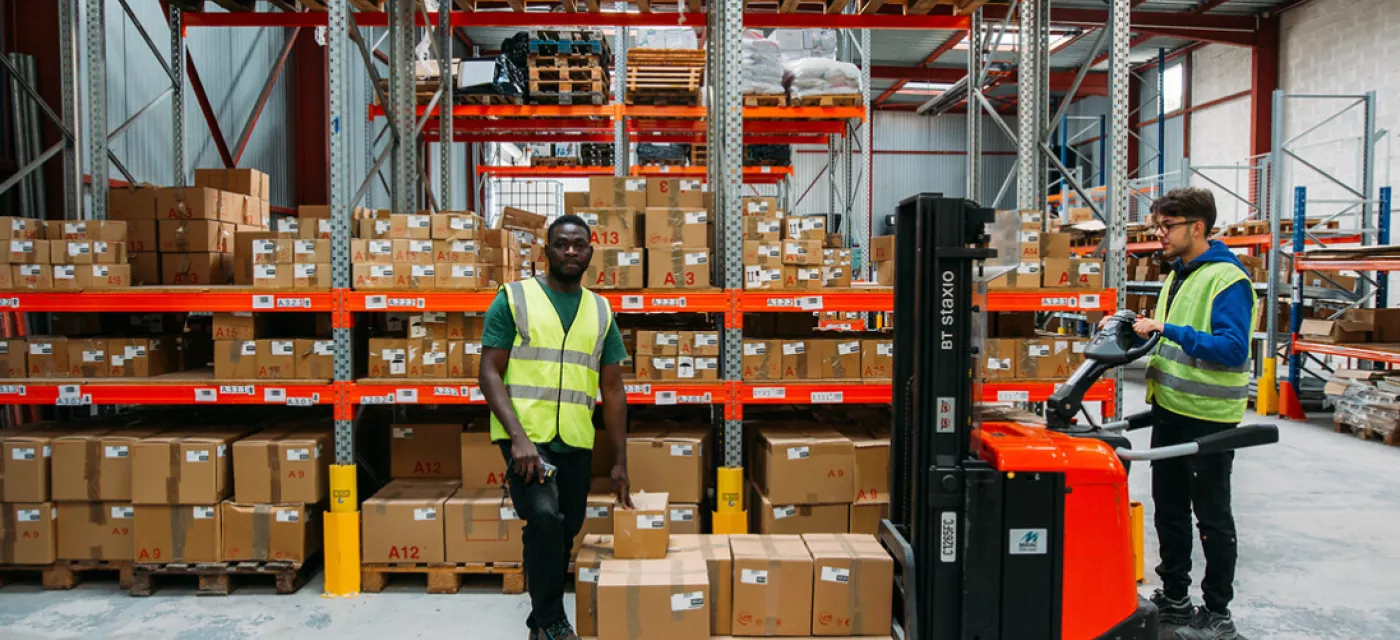 Deux jeunes dans un entrepôt déplacents de cartons à l'aide d'un transporte-palette